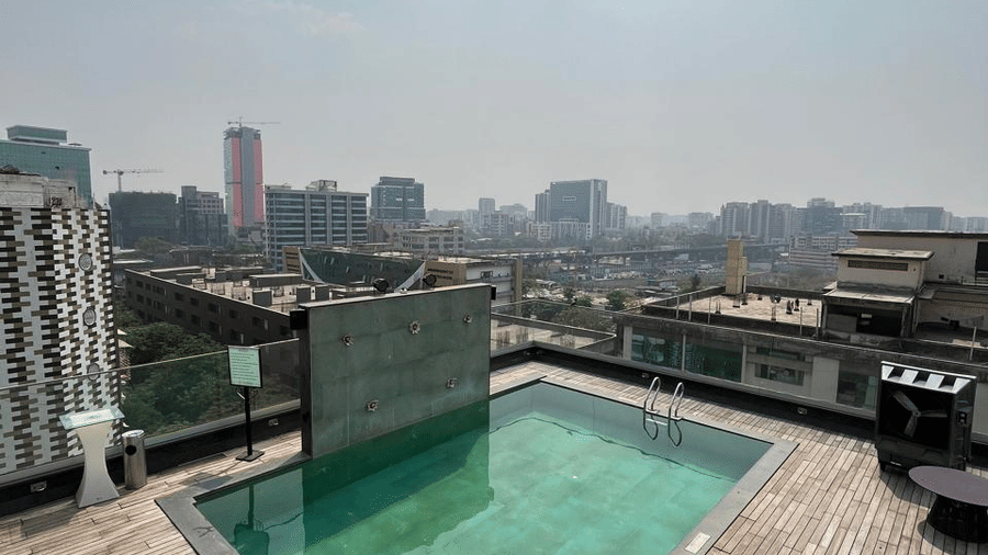 Angled view of a square rooftop swimming pool with green water on a wooden deck overlooking the city.