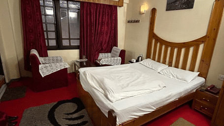 View of a bed with white linen, a wooden headboard, red carpet, and a black and white patterned rug at Hotel Dolphin Darjeeling.