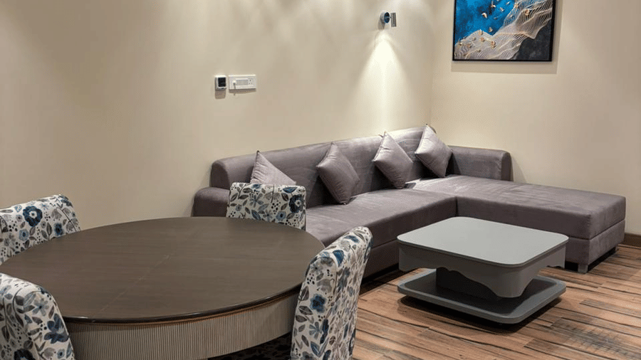 The living room with wooden flooring at the Presidential Suite at Essentia Luxury Hotel and Convention, Nagpur, featuring an L-shaped grey couch along with a dining area beside it.