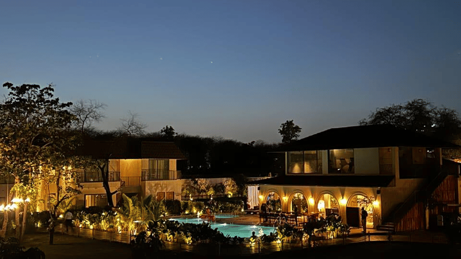 Lit swimming pool and multi storey buildings with exterior lights surrounded by dark trees under night sky at Jivana Wellness Retreat.
