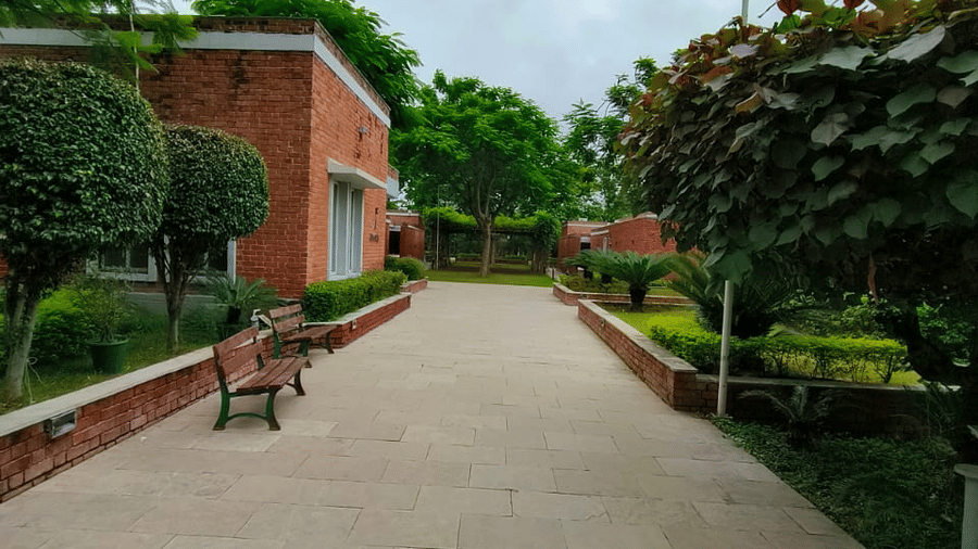 A straight, wide paved walkway leading through a grassy area towards a low, reddish-brown building under an overcast sky | Nandan Resort in Bhopal