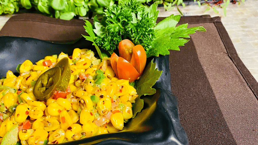 Black square plate of Mexican corn salad with yellow corn kernels, peppers and jalapenos, garnished with parsley and tomato on a brown napkin.