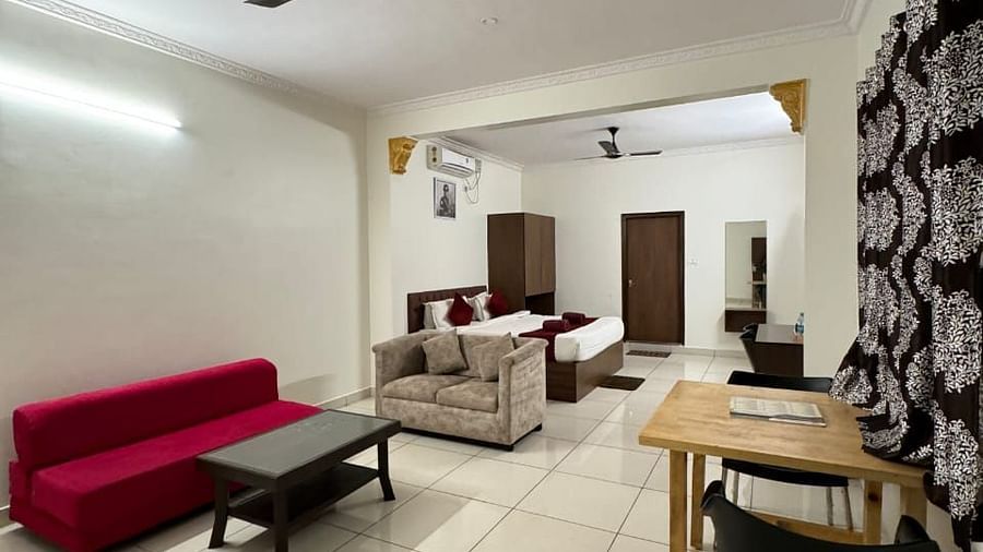 Modern living area with a beige sofa, red armchair, glass coffee table, and a plush bed in the background. | Vistar Resort, Bengaluru