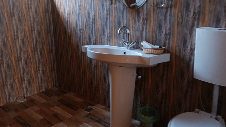 A bathroom at Dawat e Drung by FrozenFall Resort, Drung, Tangmarg, with a pedestal sink, a round wall mirror, a toilet, and wooden wall panels.