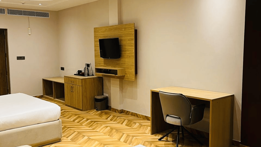 A modern hotel room with warm wooden flooring in a chevron pattern, featuring a neatly made bed, a small work desk with a chair, a mounted TV, a sofa with a white coffee table, and ceiling fans with recessed lighting - Silver Sand Beach Resort, Havelock