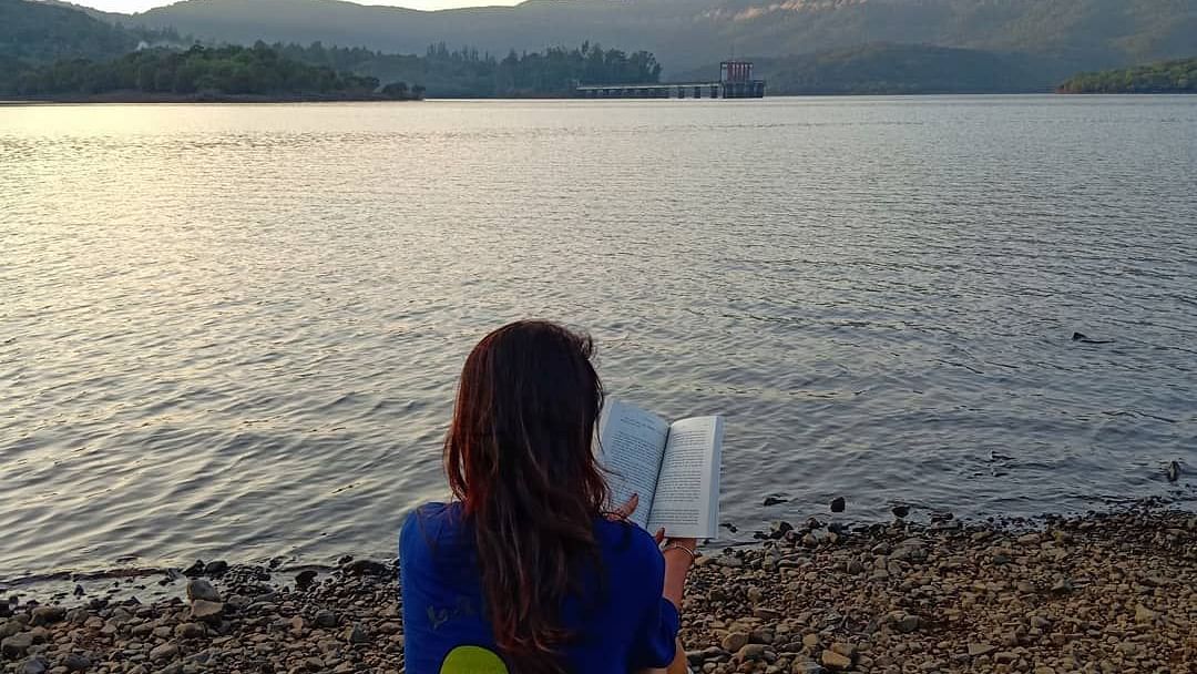 A serene view of the water and distant hills at Forest Escapes Koyna, with a person sitting peacefully by the shore.