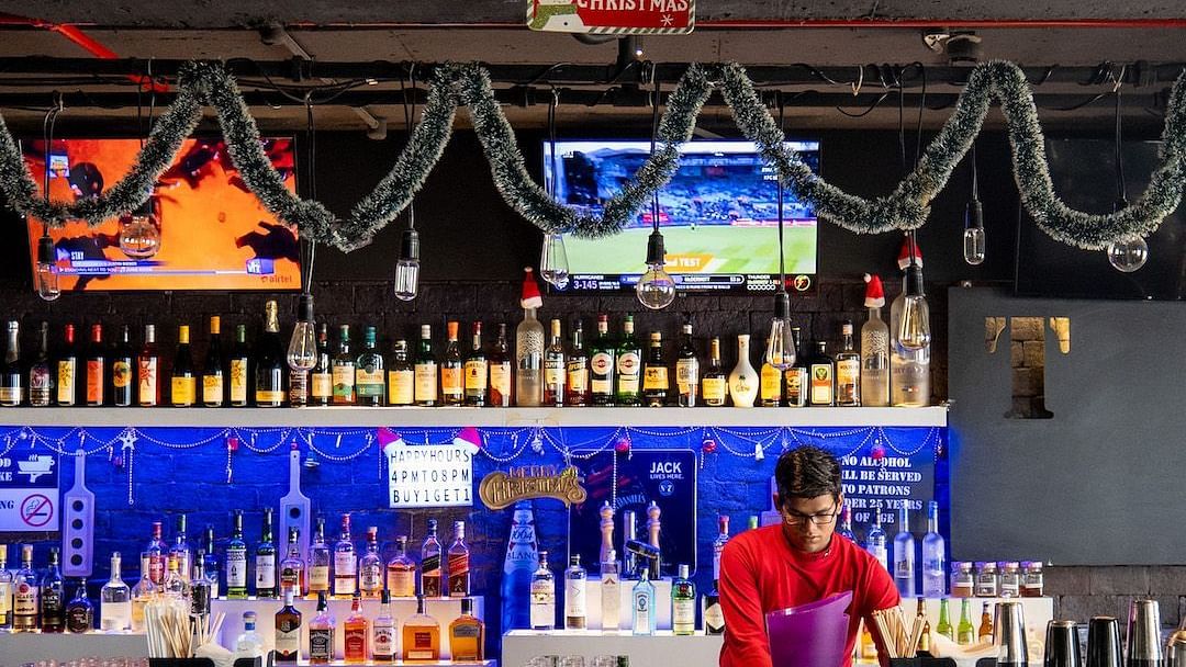 A bartender making a drink behind a brightly lit bar. The bar is decorated for Christmas with lights and garland.