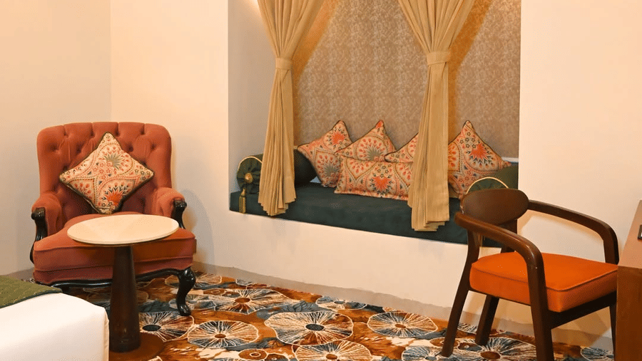 A seating area in a Kadamb Kuteer guest room with a brown armchair, a wooden desk with a chair, and large windows with decorative drapes.