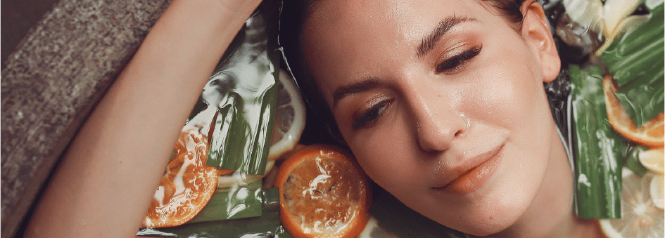 A woman in a bathtub filled with water, orange and lemon slices, and leaves, with her hands slightly lifted up