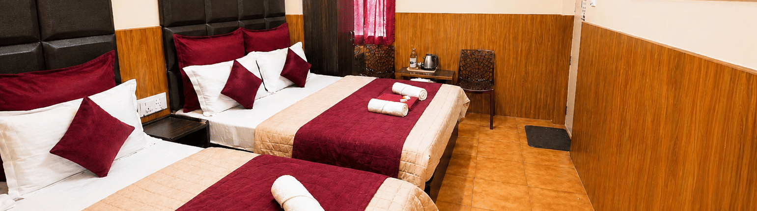 Deluxe Quadruple Room at BN Tourist Home, Ooty, featuring 2 plush beds facing a wall-mounted television screen next to a wardrobe.