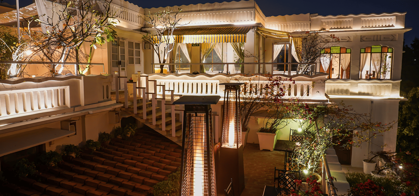 Night view of Saraca Rooftop Lounge at Saraca Hotel, Lucknow, with warm lighting, open-air seating, classic railings, and lush greenery creating an elegant terrace setting.