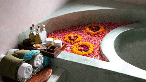 A concrete or stone bathtub filled with water and covered in red and yellow flower petals, with rolled towels and bottles on a tray nearby.