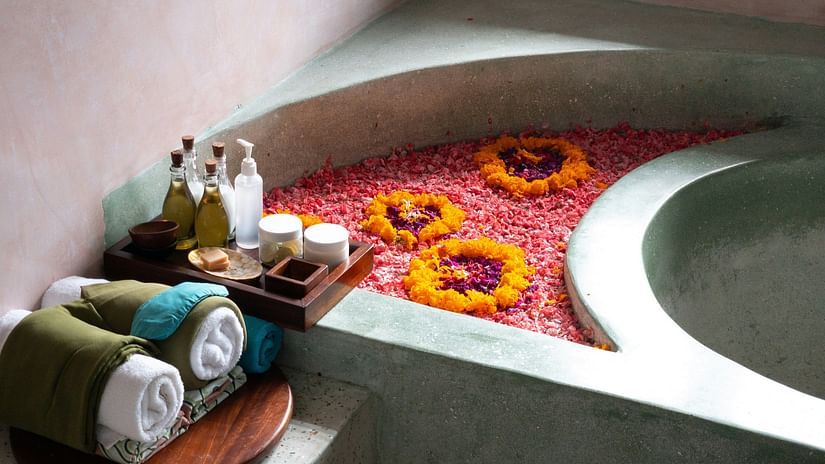 A serene spa-like scene featuring a stone or concrete bathtub filled with water and covered in colourful flower petals, with rolled towels beside it.