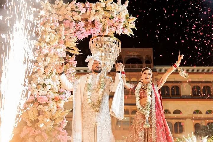 A newlywed couple celebrates under a floral arch with fireworks and petals filling the night sky, embracing the joyous moment. - Umaid Palace - A Lakeside Heritage Palace Resort