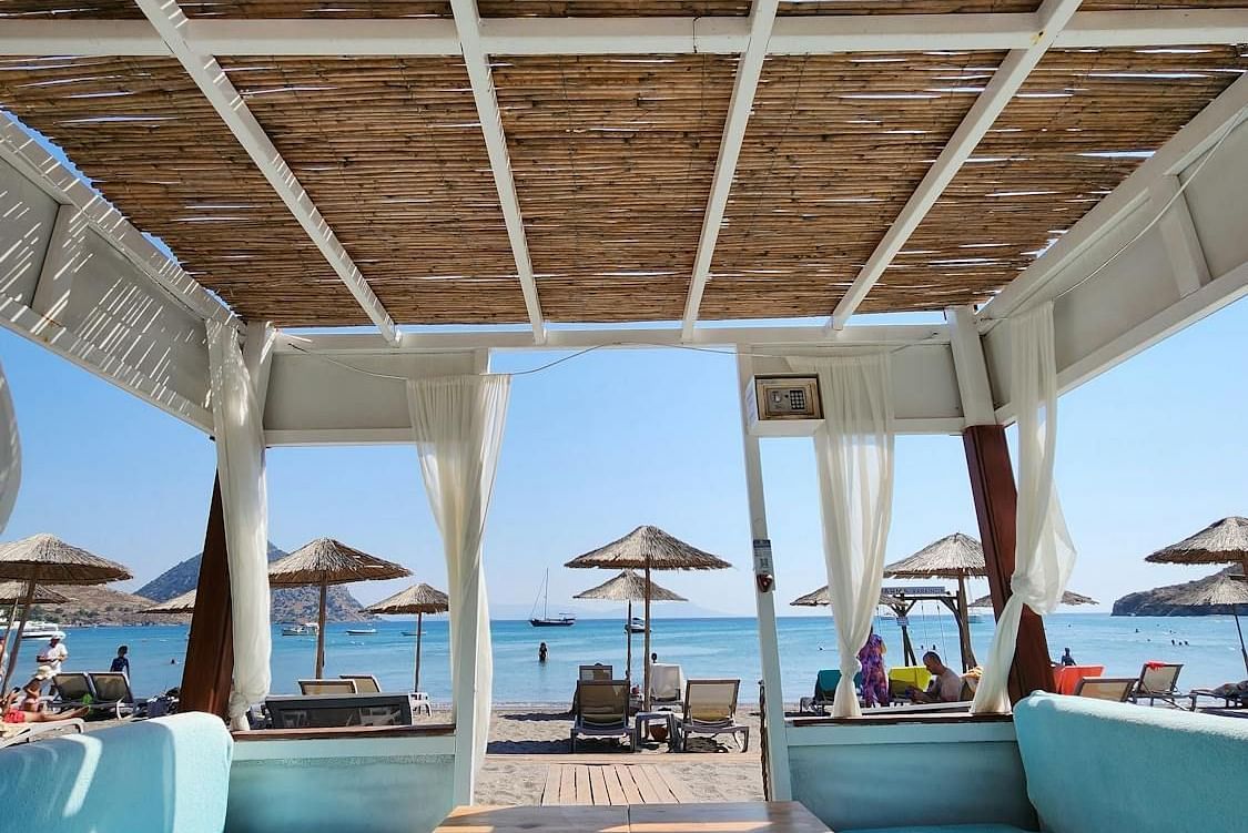 An outdoor seating area with a table and cushioned benches under a structured canopy, facing a beach scene.