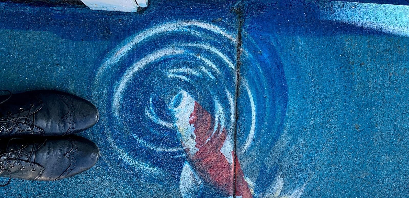 Feet in dark shoes stand next to a painted koi fish on pavement with swirling patterns.