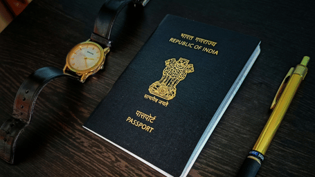 A navy blue Indian passport, a wrist watch, and a gold-coloured pen lying on a dark wooden surface.