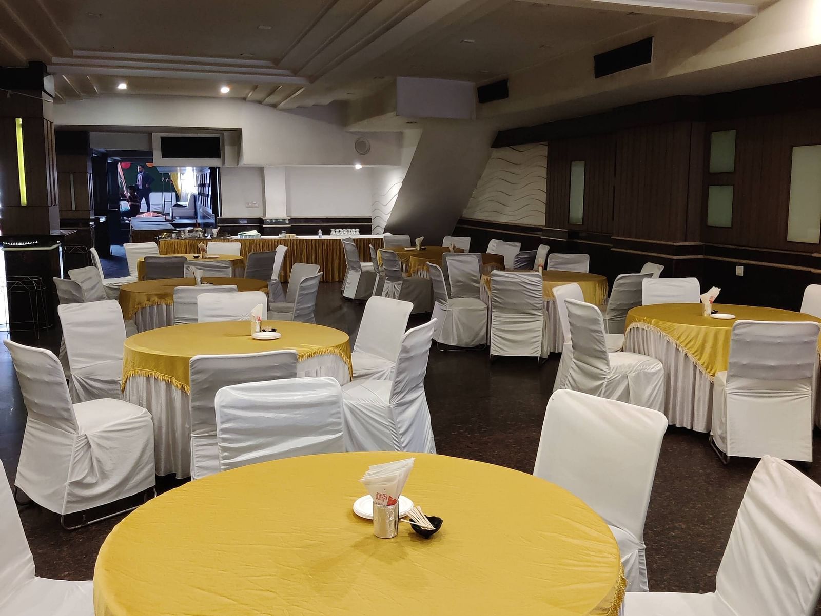 A few round tables covered with bright yellow coloured table cloth and chairs surrounding it with a stage at the far end of the Banquet Hall at Hotel Gold, Panipat.