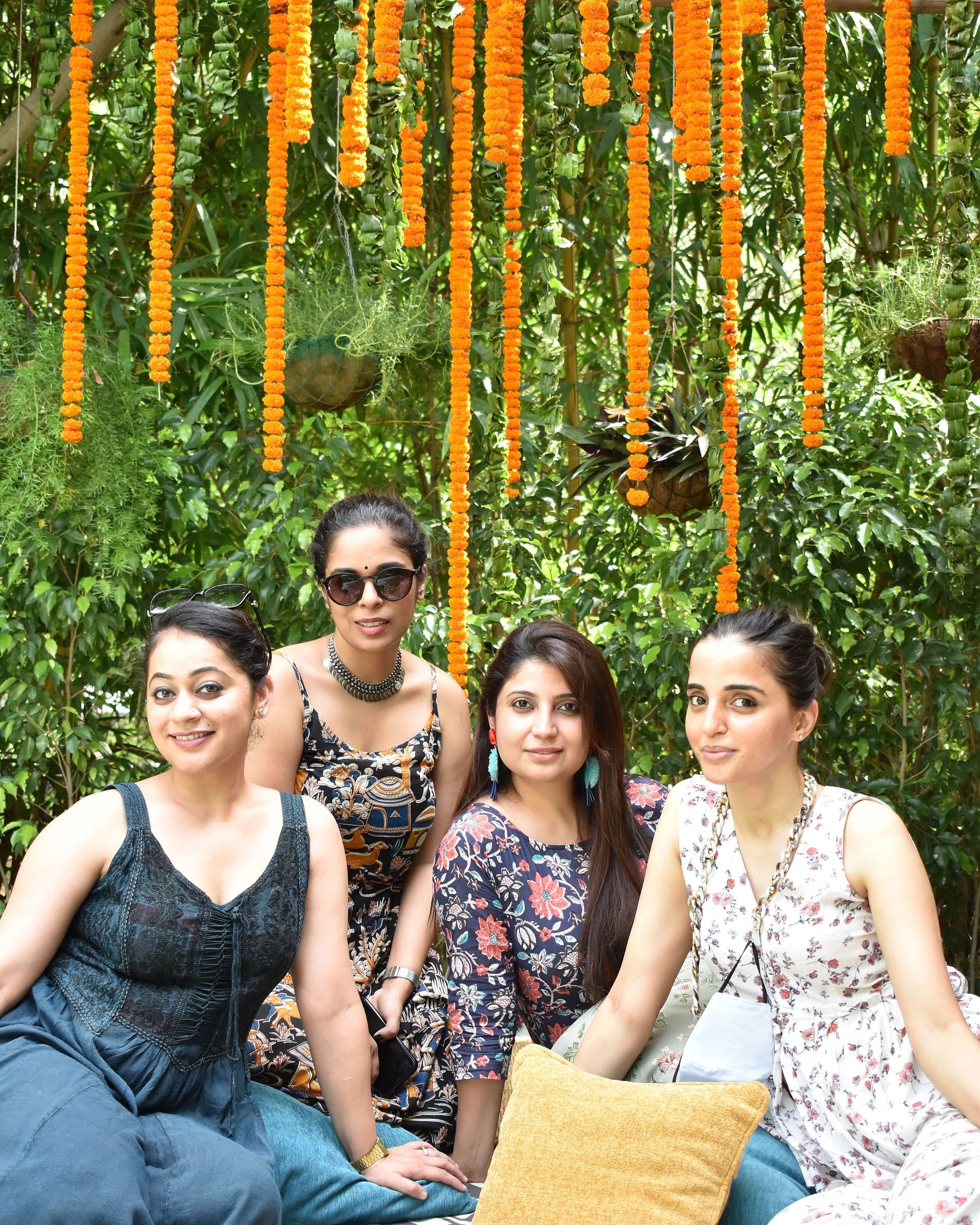 Four people sitting on a cloth with flowers hanging for overhead decorations and a background that's covered with plants and trees at Karma Chalets.