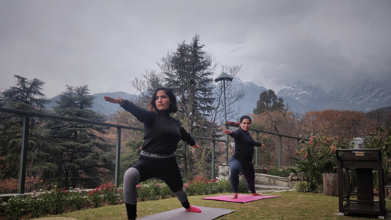 Two women practicing yoga outdoors, one in warrior pose and the other in tree pose, with mountains in the background.