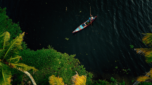 A small, colourful boat with a person paddling on dark water, bordered by lush green tropical vegetation.