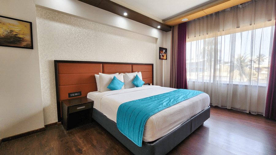 A spacious room at Dimora, Thiruvananthapuram, featuring a well-made bed, a headboard, a bedside table, and daylight streaming in through sheer curtains.