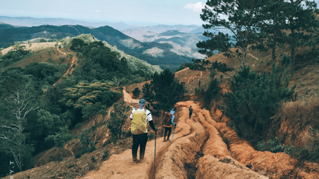 A person walking along a dirt trail on a hillside with trees and valleys visible in the background.