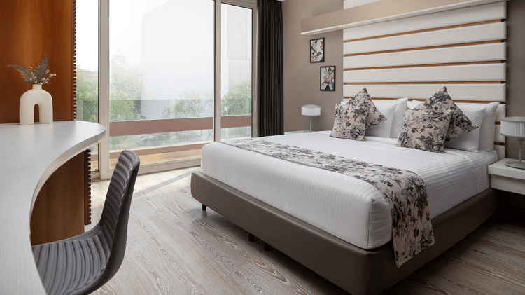A bright room at The Atara, Gurugram boasting a large window offering an outdoor view, a comfortable bed with a floral runner, and a minimalist white desk with a chair