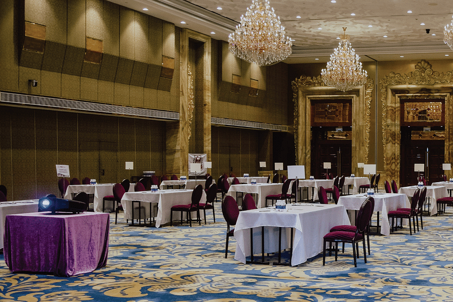 A grand ballroom features elegant chandeliers, set tables with maroon chairs, and a projector on a covered table with text - Opening Soon in Bichpuri Agra.