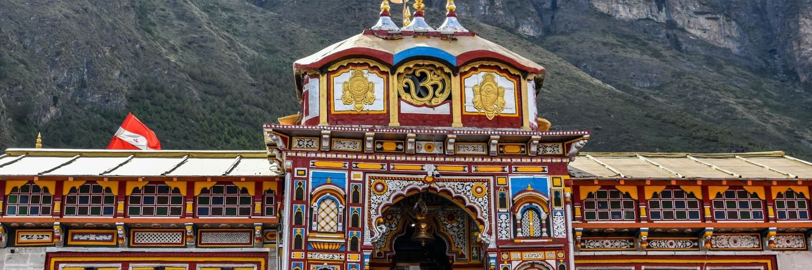 Crowd entering colourful temple with flags, set against towering mountain cliffs under blue sky, a must-visit while staying at Badrinath Hotels Near Temple