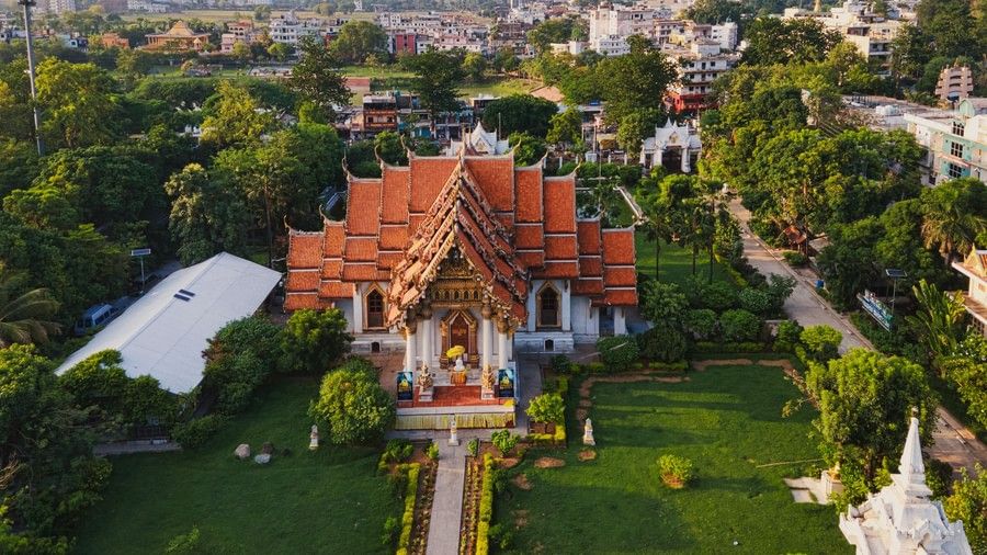 Aerial view of a traditional temple with ornate red-tiled roofs and golden details, surrounded by lush green gardens and pathways, set against a backdrop of a bustling cityscape.