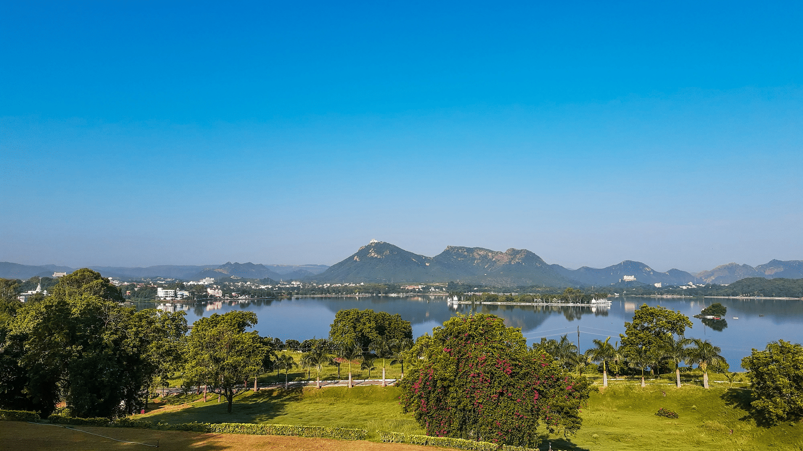 An aerial view of the Fateh Sagar Lake with its surrounding greenery and mountains under a bright blue sky.