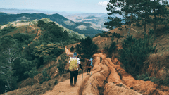 2 people walking on a dirt path along a hillside, with trees on one side and layered hills and valleys visible in the distance.