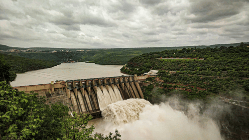 An overview of Aliyar Dam, Pollachi, with water gushing out from the vents, with the dam surrounded by trees under a cloudy sky