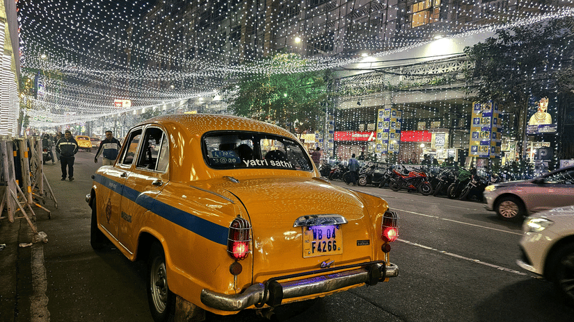 A cab parked on the side of Park Street with other vehicles moving behind it and the street decorated with lights in view.