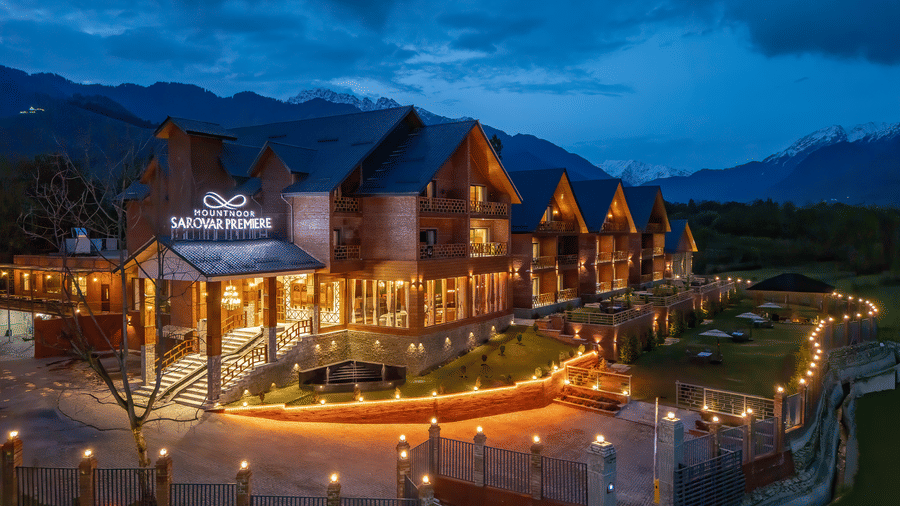 Facade of Mountnoor Sarovar Premiere resort in Pahalgam, with warm lights and the perimeter fence glow against the deep blue dusk sky, highlighting the traditional mountain-lodge aesthetic.