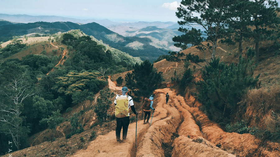 2 people walking on a dirt path along a hillside, with trees on one side and layered hills and valleys visible in the distance.