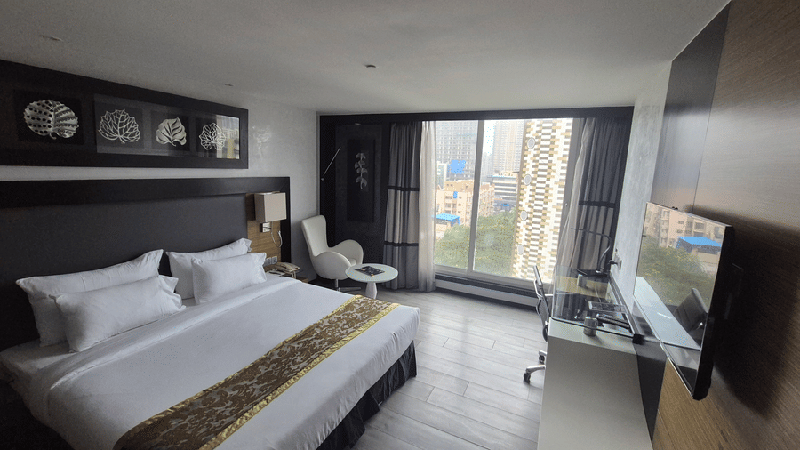 Spacious contemporary hotel room with a double bed, wooden floors, and a large window looking out at high-rise buildings.