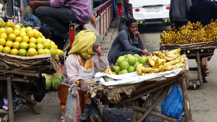 Street vendors sitting on the side of the street selling fruits and vegetable in Mercato Market with people walking by in the background.