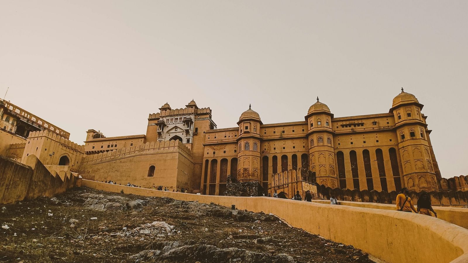 A wide shot of Amber Fort with yellow sandstone walls and ramparts against a pale sky, with a rough foreground