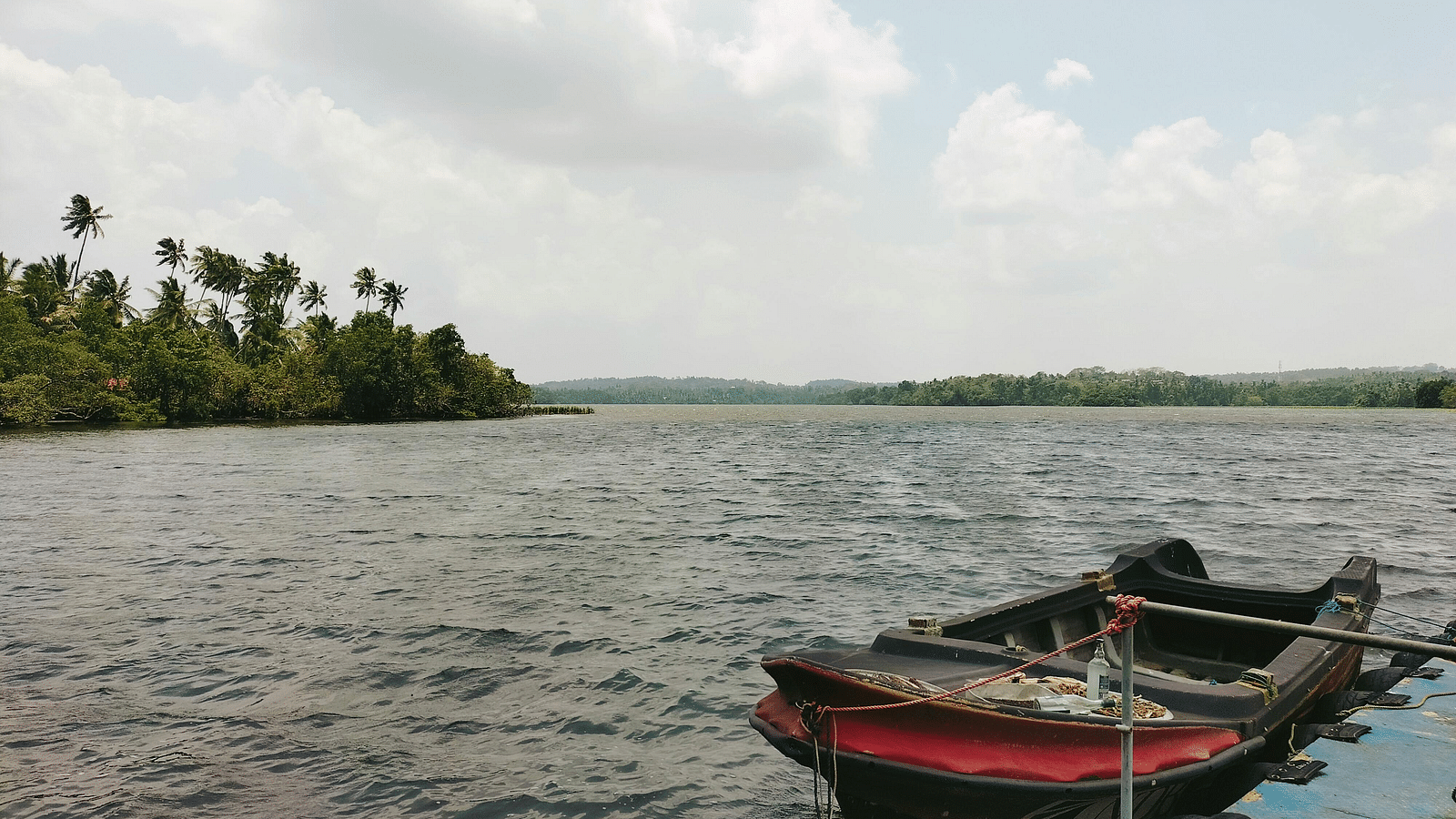A wooden boat anchored on the calm waters of a lake, with a backdrop of lush green trees and a cloudy sky in the distance