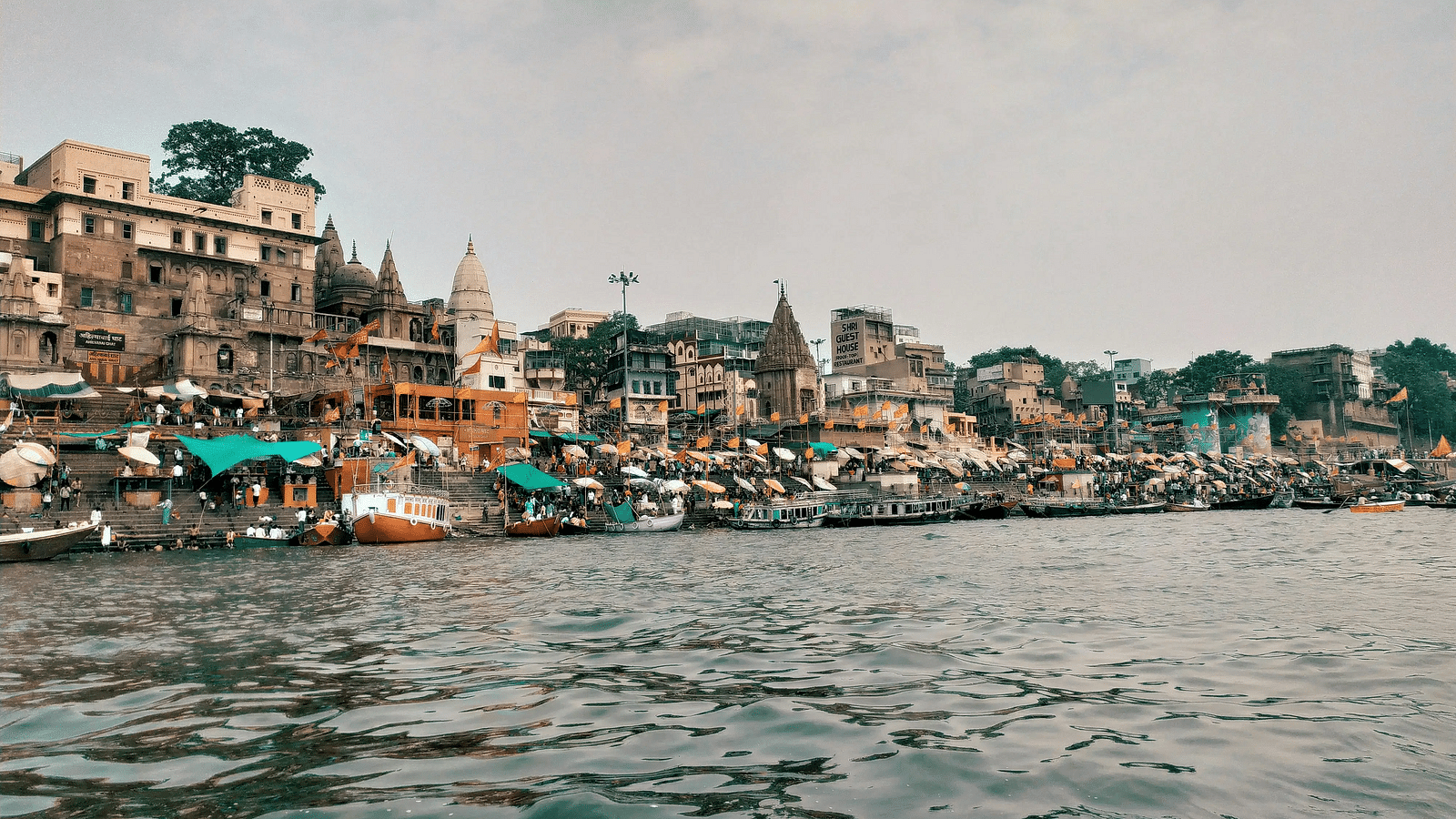 A wide view of Varanasi ghats from the Ganges River with historic buildings in the background.