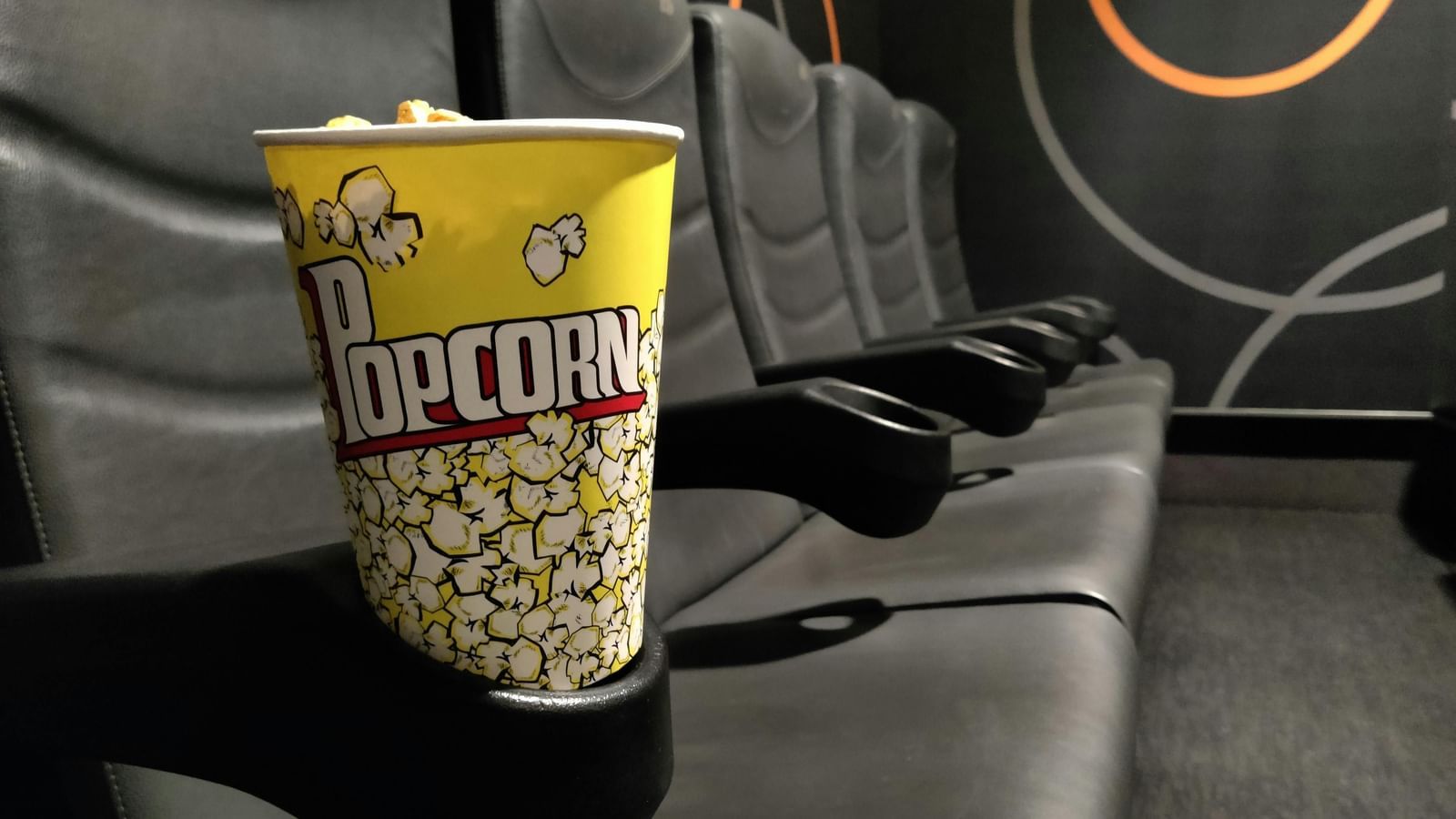 A yellow popcorn bucket placed on a black cinema seat inside a movie hall.