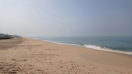 A wide, serene sandy beach meets the gentle waves of the sea under a hazy blue sky.