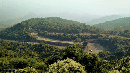 A car travelling through scenic winding ghat road cutting through lush green hills.
