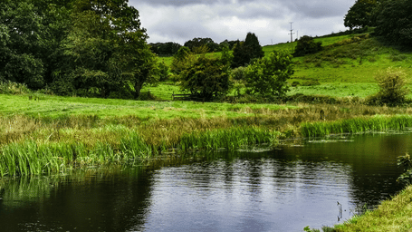A tranquil river flows through a lush green valley with rolling hills under a cloudy, overcast British sky.