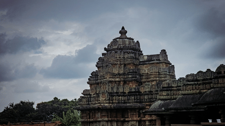 Historic stone temple tower rises beneath a cloudy sky, with intricate carvings and weathered masonry surrounding its ancient architectural form.