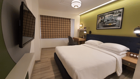 A modern and comfortable hotel room with a king size bed, a bedside table, a work desk with a chair, a  TV, and a window with curtains  | T24 Retro