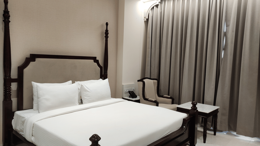 A bedroom at Anant Mahal, Jaipur featuring a king size bed with multiple pillows arranged at the head of the bed, a patterned rug, floor to ceiling curtains behind a table, and a chair.