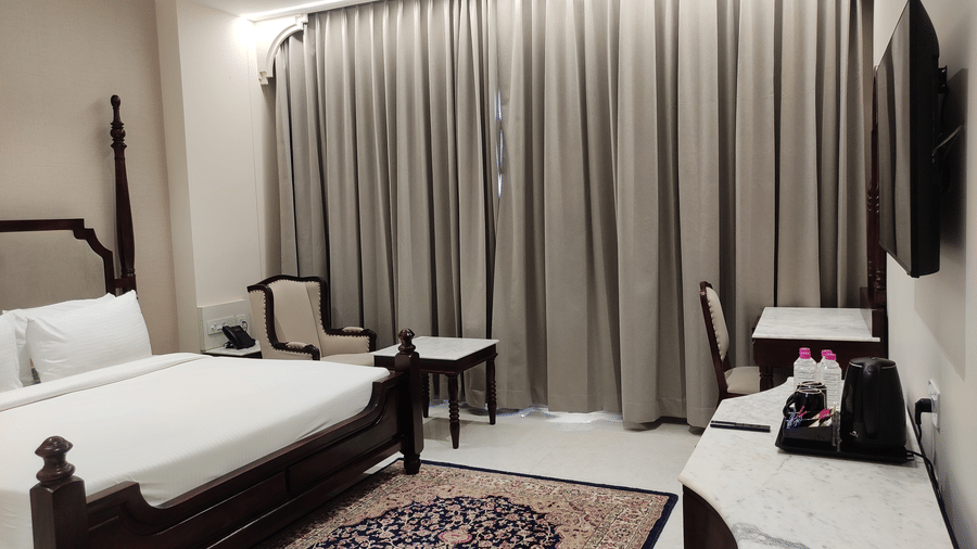 A room interior at Anant Mahal, Jaipur featuring a king size bed, a patterned rug on the floor, a wall mounted television, and long curtains covering a large window behind the seating area.